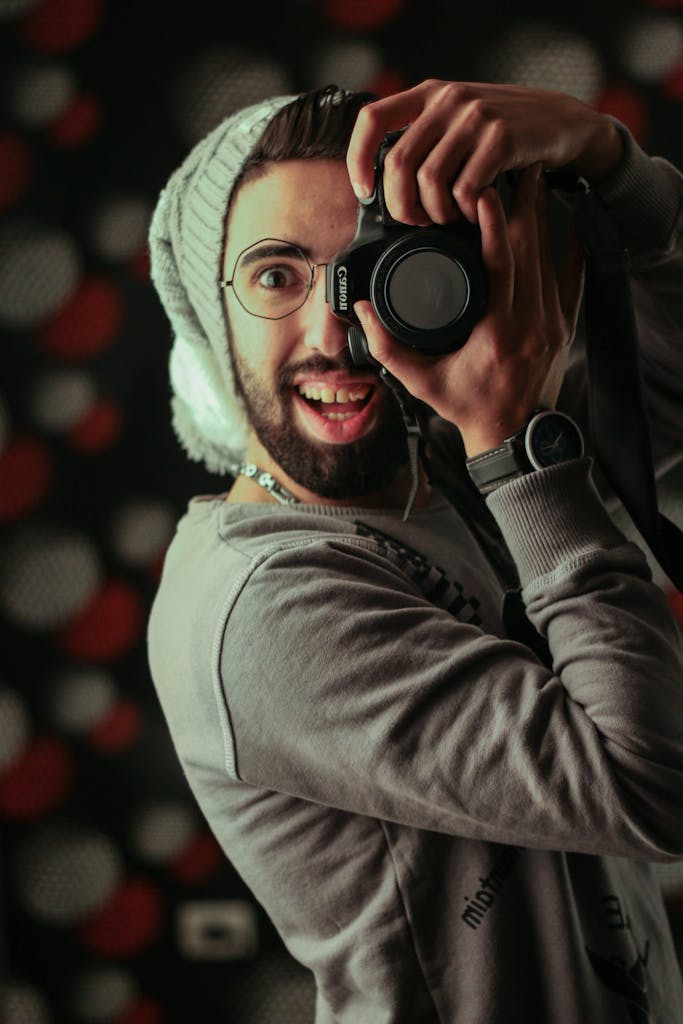A playful male photographer in a beanie and eyeglasses takes a creative selfie with a DSLR in a studio setting.